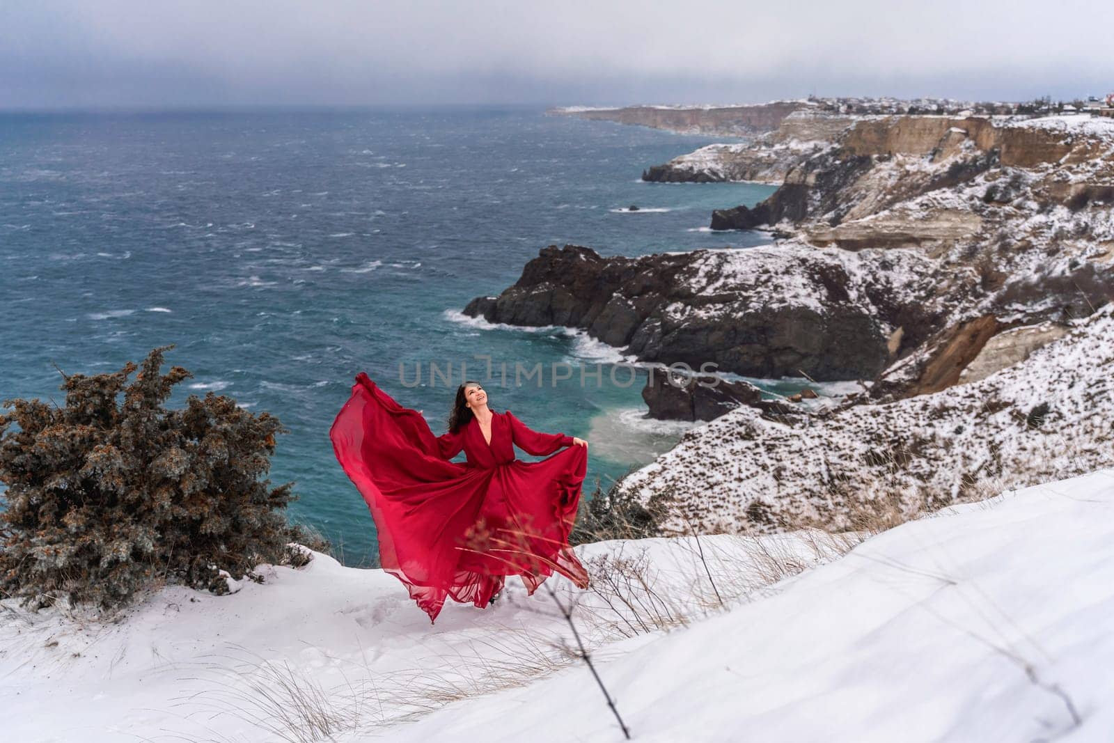 Woman red dress snow sea. Happy woman in a red dress in the snowy mountains by the emerald sea. The wind blows her clothes, posing against sea and snow background