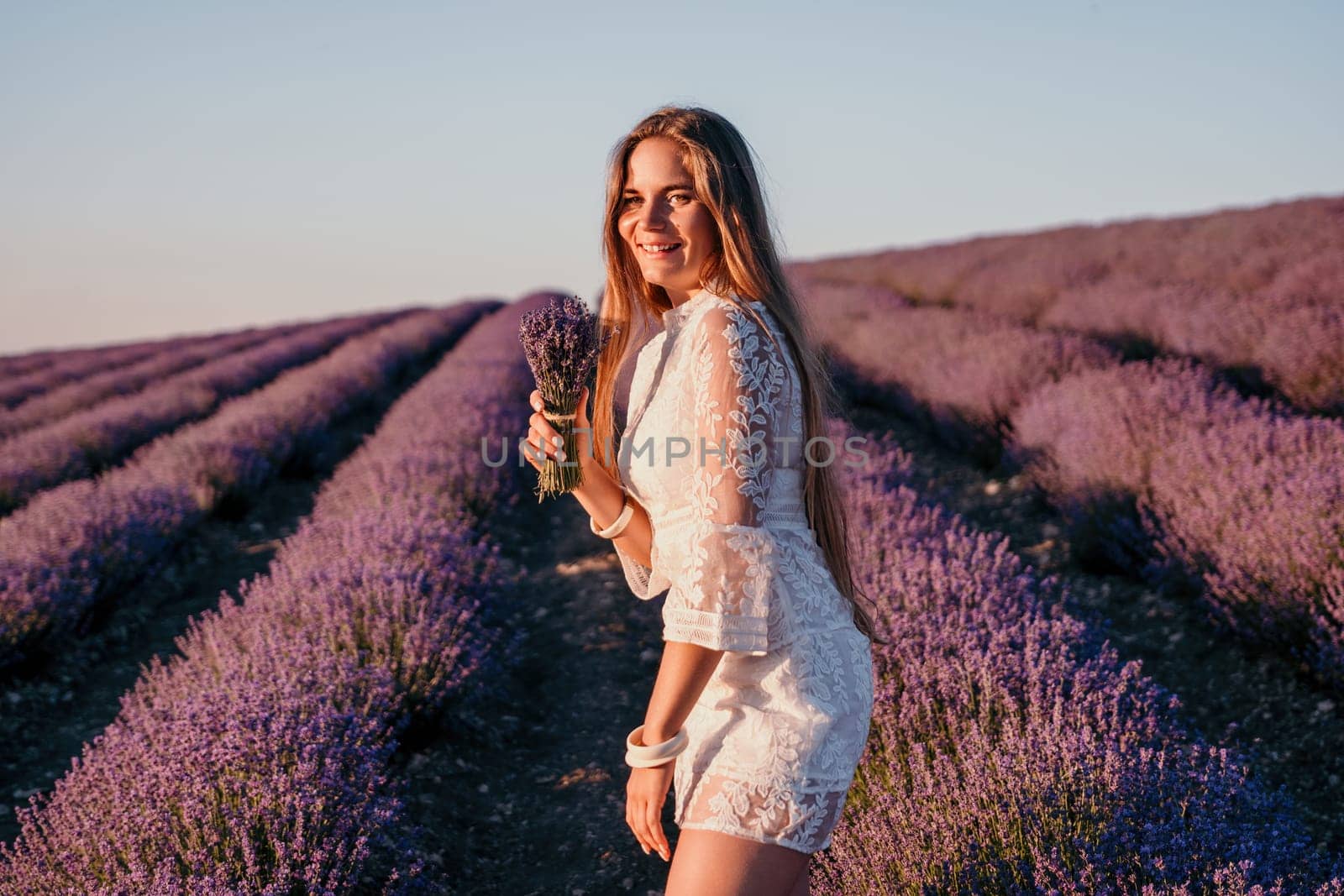 Close up portrait of young beautiful woman in a white dress and a hat is walking in the lavender field and smelling lavender bouquet.