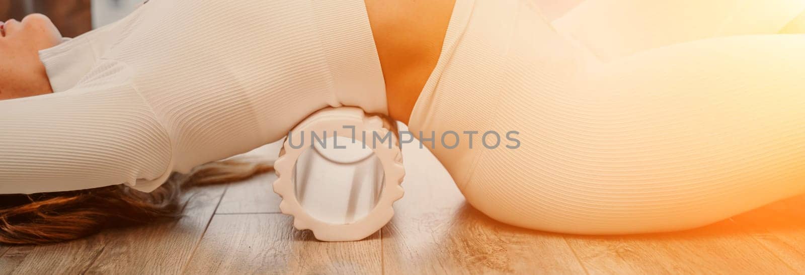 Adult athletic woman, in white bodysuit, performing fascia exercises on the floor - caucasian woman using a massage foam roller - a tool to relieve tension in the back and relieve muscle pain - the concept of physiotherapy and stretching training by Matiunina