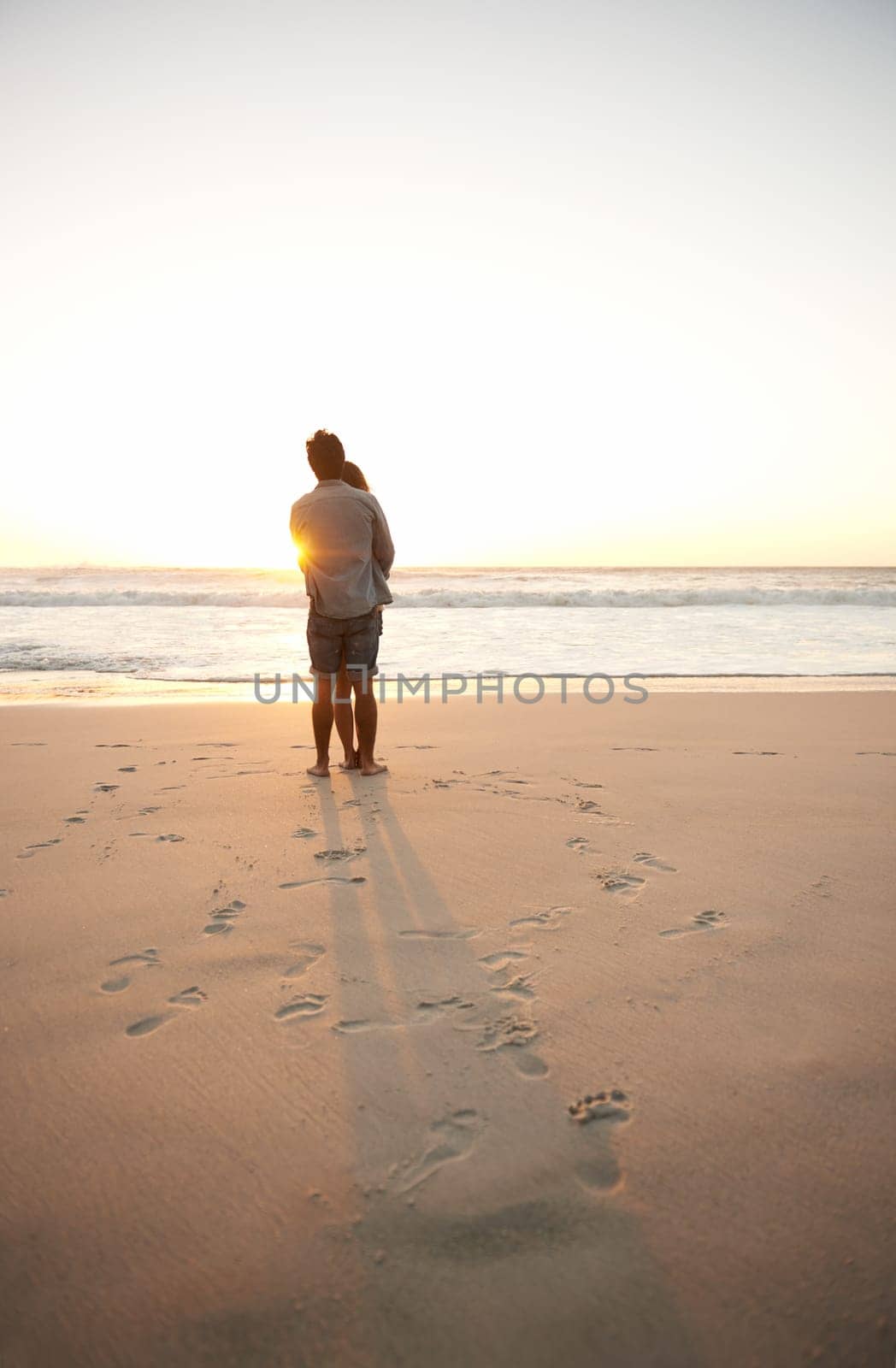Man, woman and beach love at sunset for romantic summer travel or adventure, bonding or marriage. Couple, footprints and embrace at water for dating tourism or nature vacation, environment or holiday.