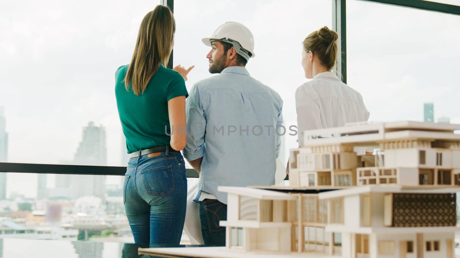 Group of caucasian diverse architect engineer pointing at skyscraper at day time. Skilled manager team looking at city or skyscraper while house model was placed on table. Back view. Tracery