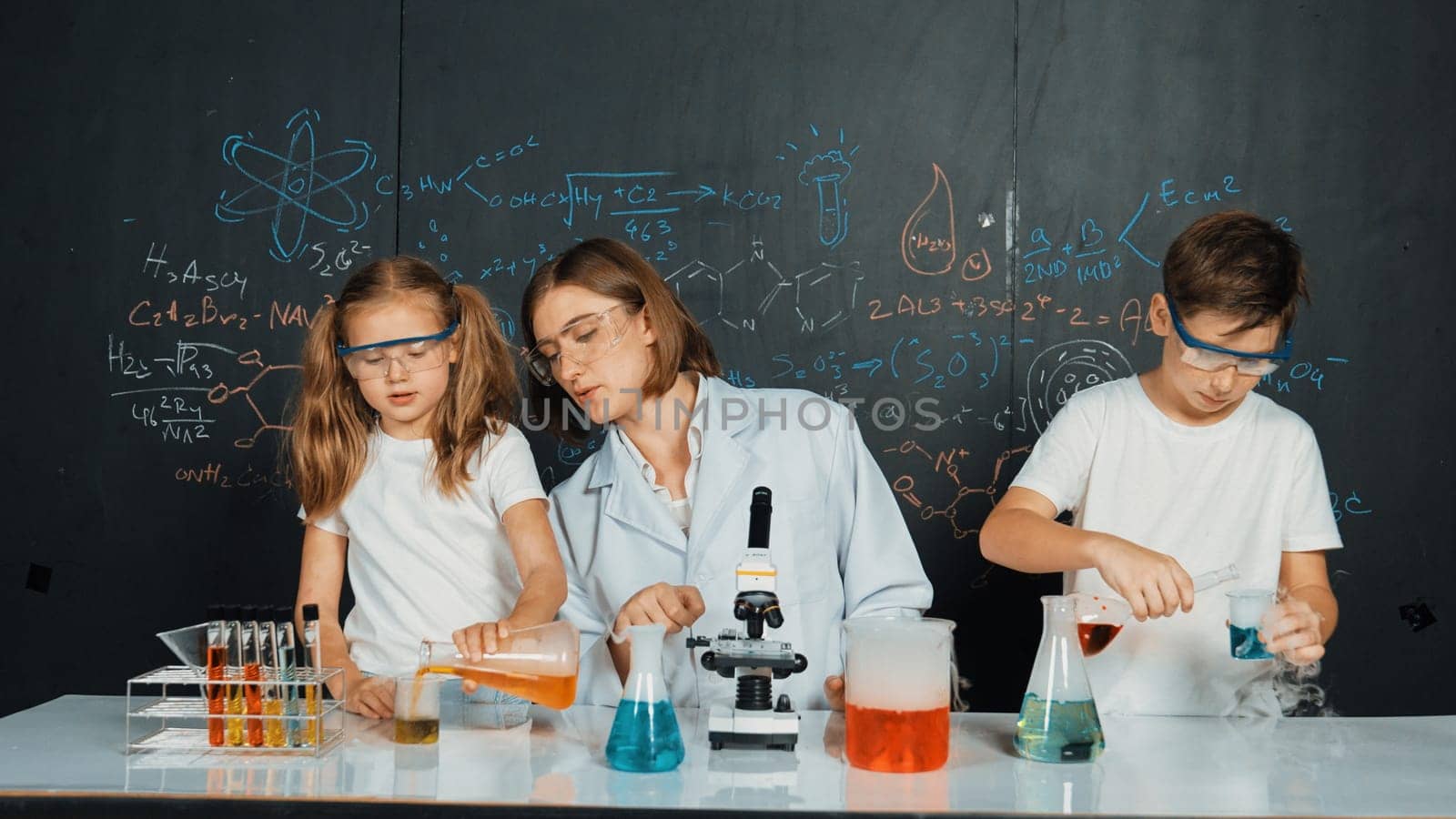 Caucasian boy mixing chemical liquid while teacher giving advice. Professional instructor wearing lab suit looking for diverse student at table with beaker filled with colored solution. Erudition.