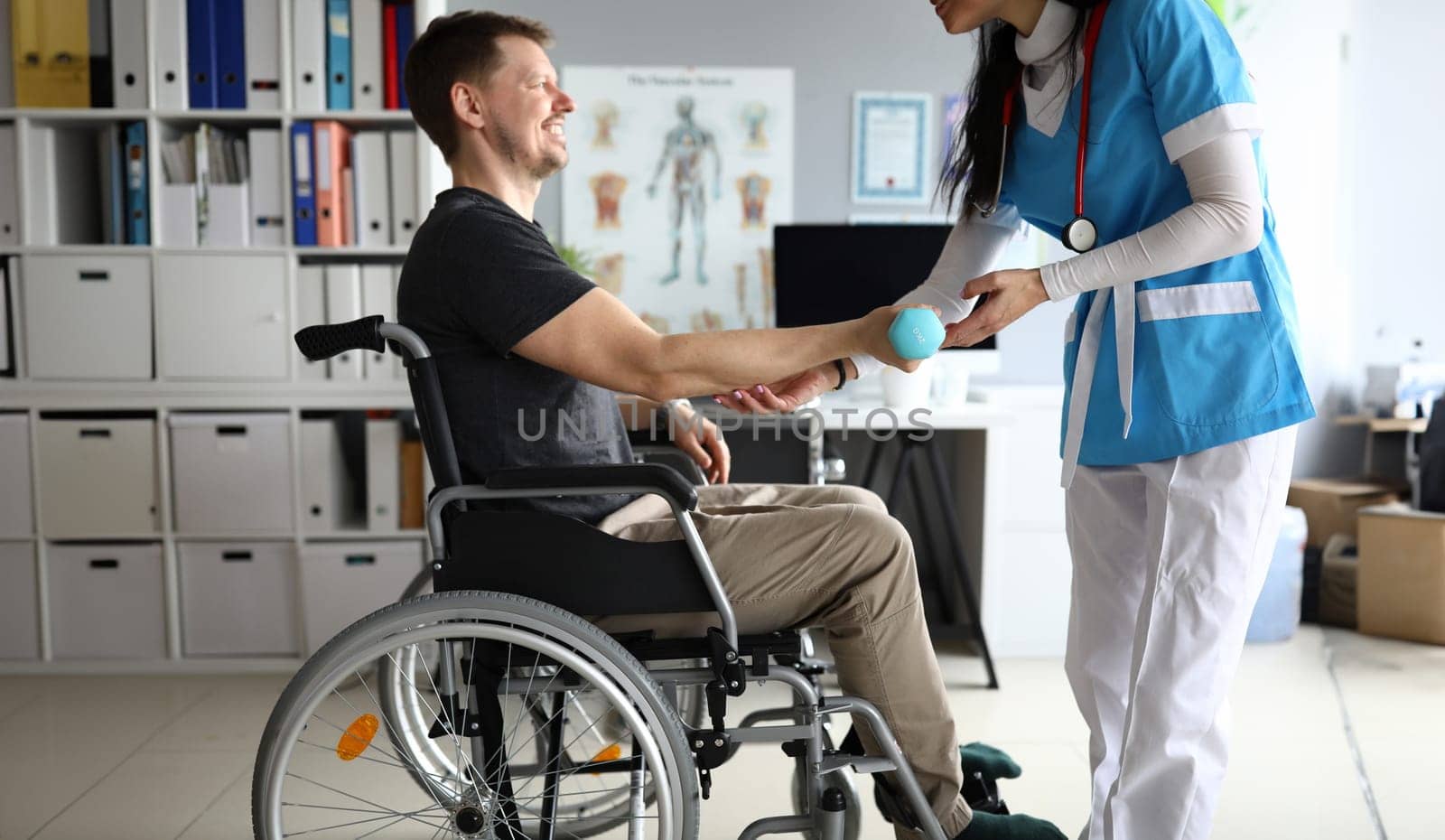 Doctor helps man on wheelchair hold dumbbell. Strengthening human parts, ability to part with wheelchair. Exercise is suitable for people with disabilities who have strength in their upper body