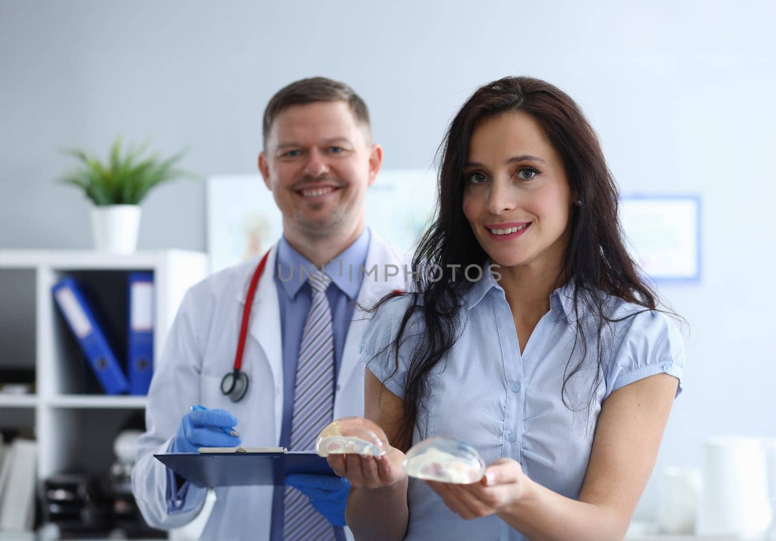 Girl in clinic holds breast implants near doctor. Comfortable atmosphere. Work plastic surgeon. Selection size breast prostheses. With men plastic surgeon at preoperative consultation