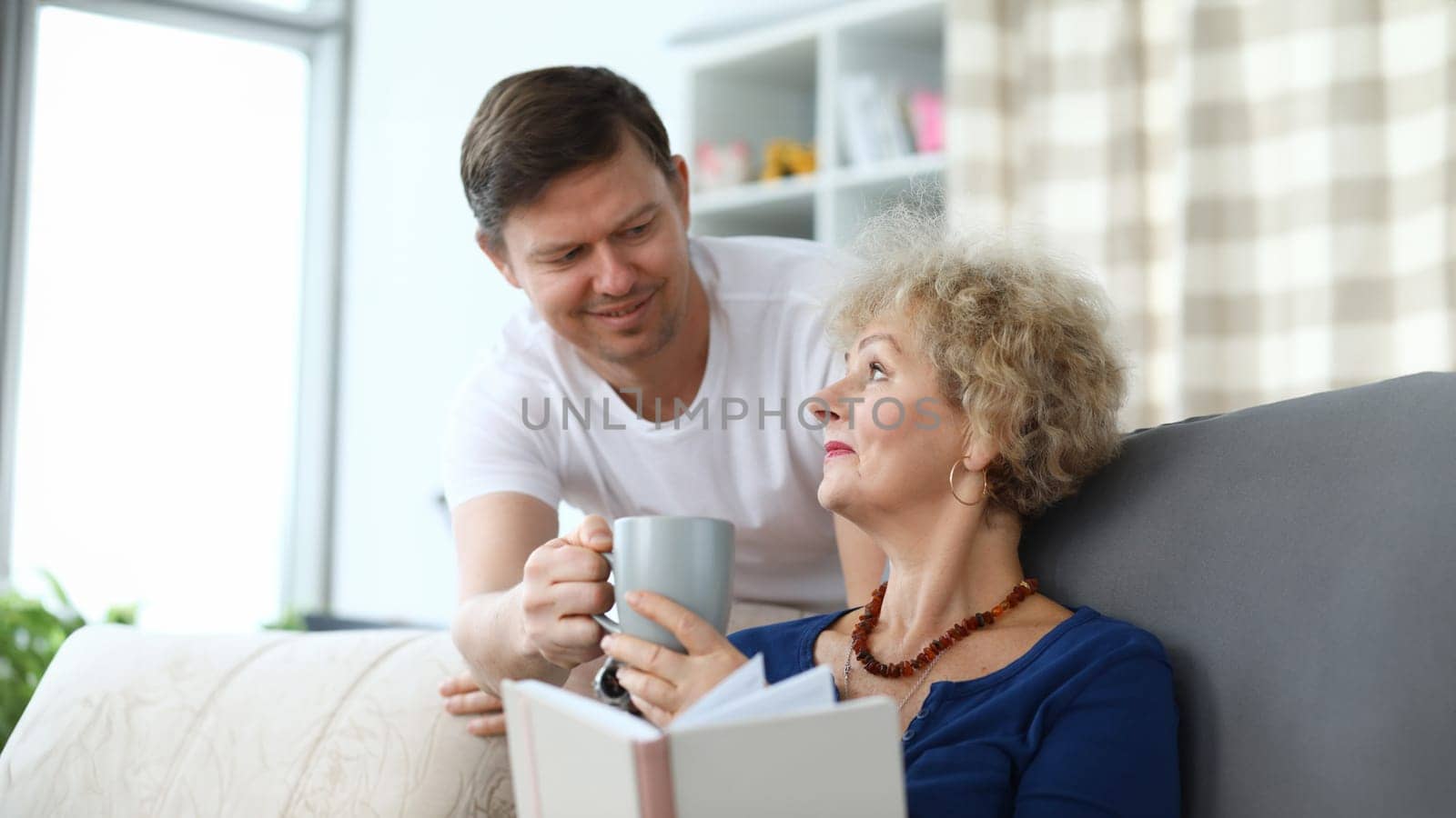 Adult son serves cup tea to an elderly mother. Relatives help each other during self-isolation. Time for reading fiction and books on self-development. Son pays attention to an elderly mother