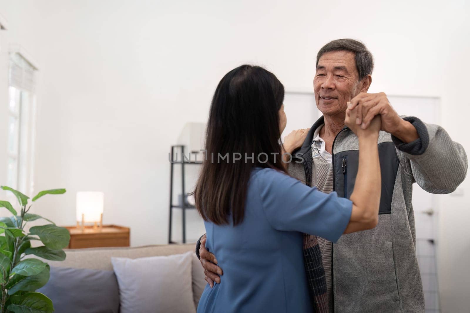 Elderly couple in love looking at each other with tenderness while dancing together in living room at home.