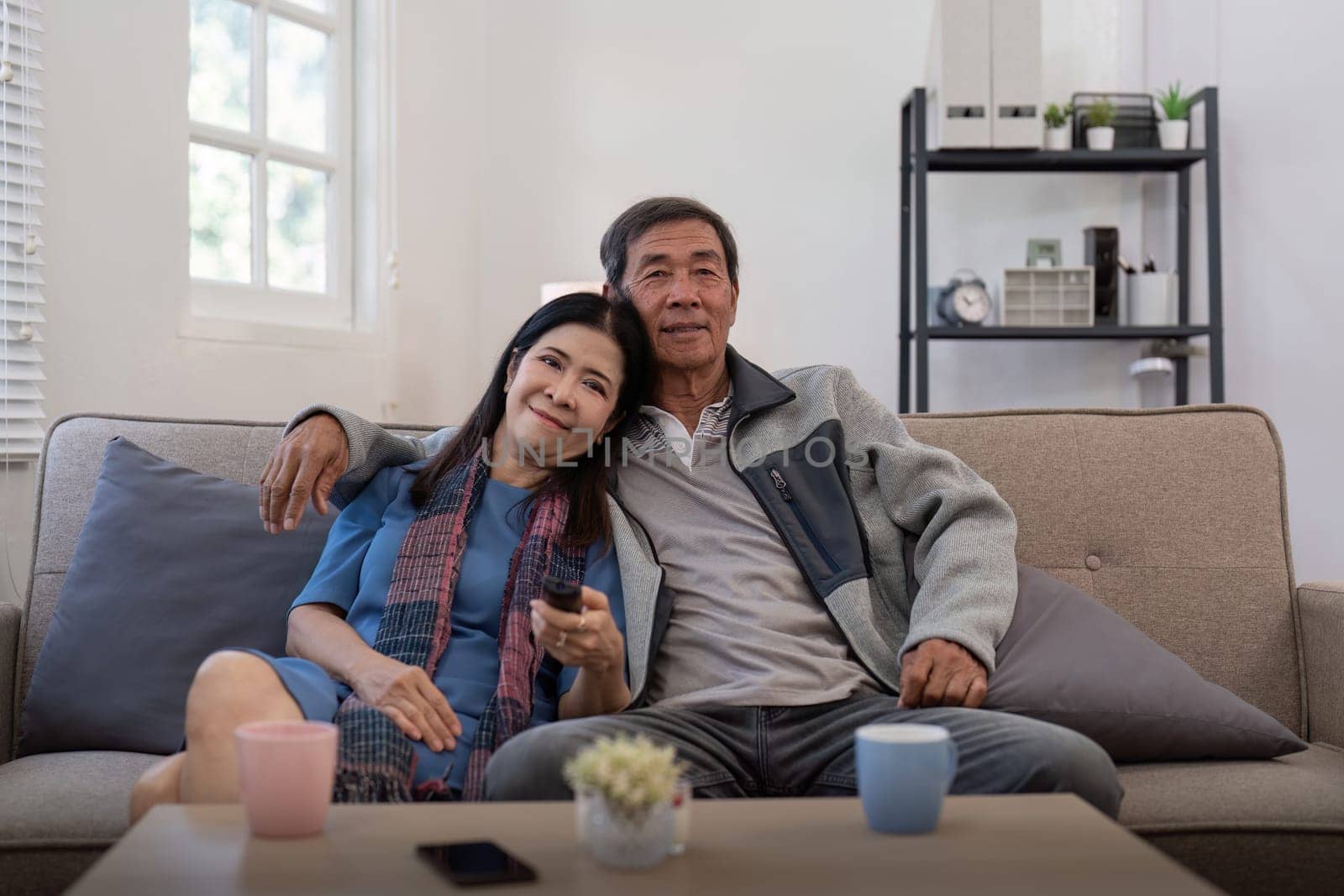 Senior couple watching tv and sofa in relax for movie or series in living room at home. Elderly man and woman with coffee and remote together for changing channel and online entertainment by nateemee