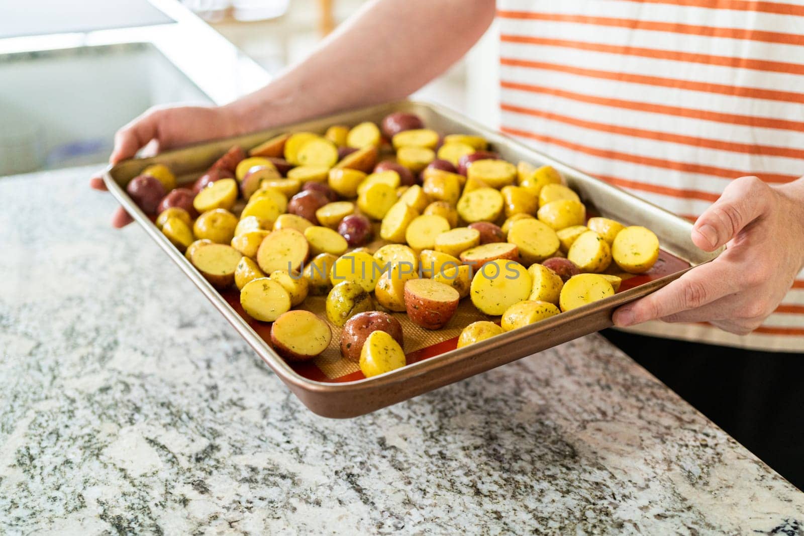 In a modern kitchen, an array of halved, multicolored marble potatoes are arranged on a baking pan lined with a silicone liner. The roasting process infuses the kitchen with a mouthwatering aroma, indicating a delicious side dish in the making.