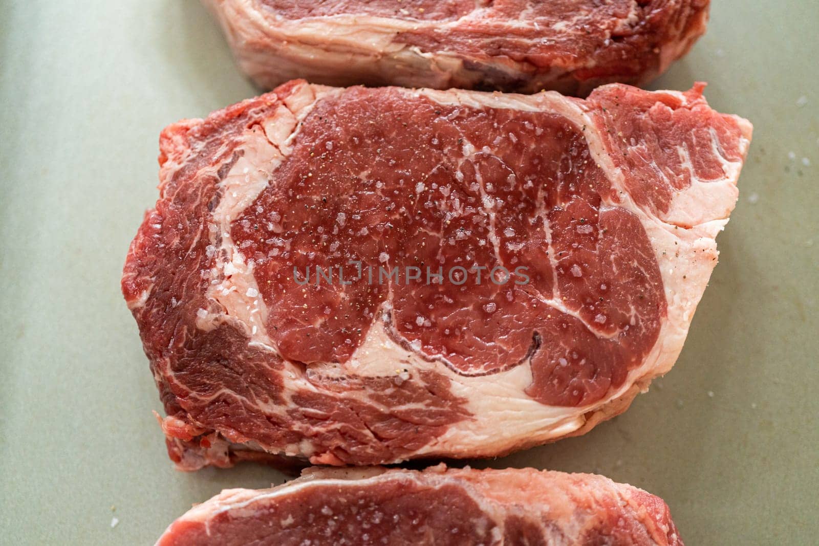 Situated in a modern white kitchen, a seasoned rib eye steak, boasting its beautiful marbling, sits ready on a baking sheet. It is prepared for the outdoor gas grill, promising a perfect sear on the exterior and a juicy, tender interior, heralding an upcoming indulgent feast.