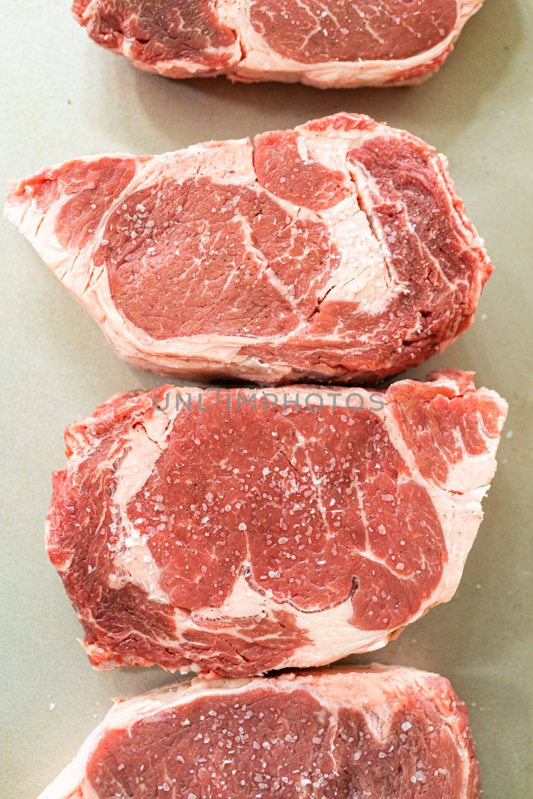 Situated in a modern white kitchen, a seasoned rib eye steak, boasting its beautiful marbling, sits ready on a baking sheet. It is prepared for the outdoor gas grill, promising a perfect sear on the exterior and a juicy, tender interior, heralding an upcoming indulgent feast.