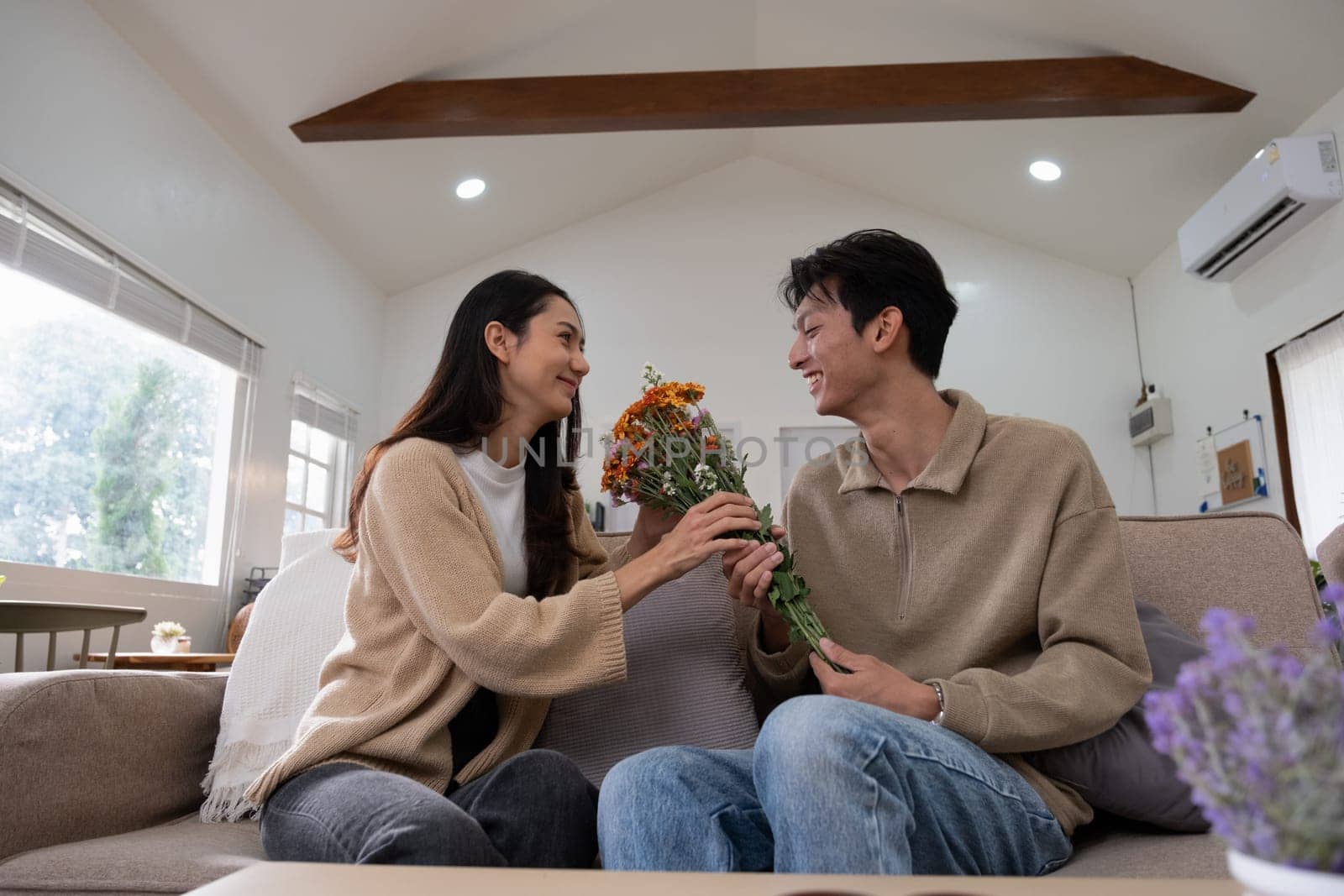 Romantic young asian couple embracing with holding flowers and smiling in living room at home. fall in love. Valentine concept by wichayada