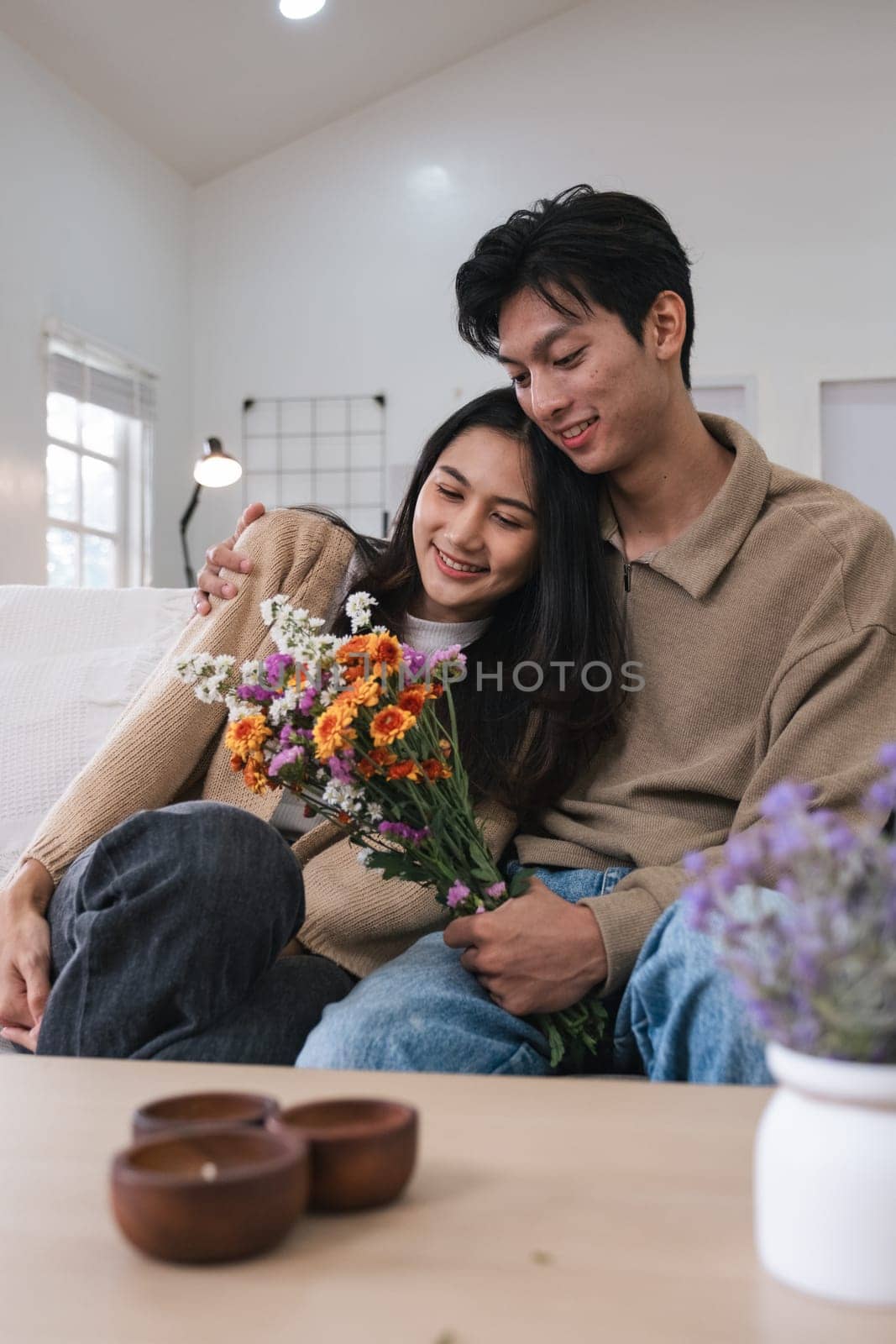 Romantic young asian couple embracing with holding flowers and smiling in living room at home. fall in love. Valentine concept.