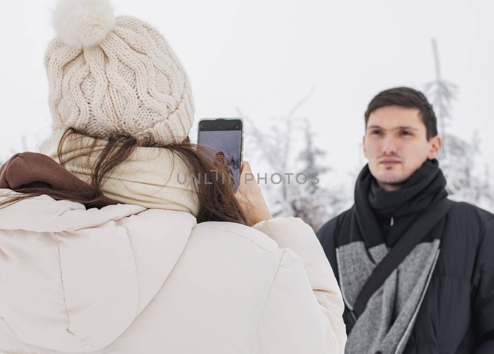 A young girl in winter clothes takes a picture on a smartphone of her handsome caucasian brunette boyfriend in a blue down jacket standing in a winter forest reserve in belgium, close-up side view. Concept of winter holidays, using technology.