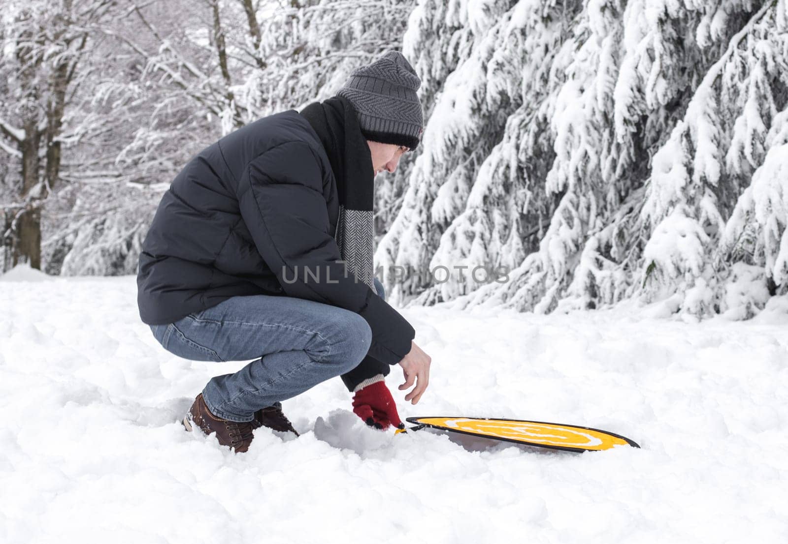 A young man in a blue down jacket and a gray knitted hat squatting sets up a yellow pad for a drone on a snowy meadow in a winter forest reserve in belgium, close-up side view. The concept of winter holidays, using technology.