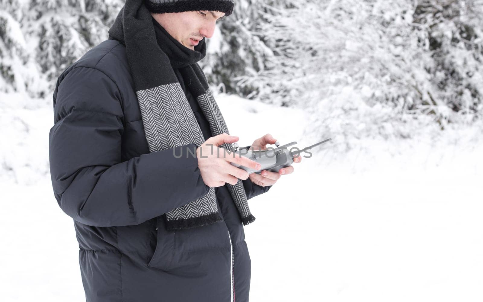 A young handsome caucasian man in a blue down jacket and a gray knitted hat controls a drone through a gray remote control in his hands while standing on a snowy meadow in a winter forest reserve in Belgium, close-up side view. The concept of winter holidays, using technology.