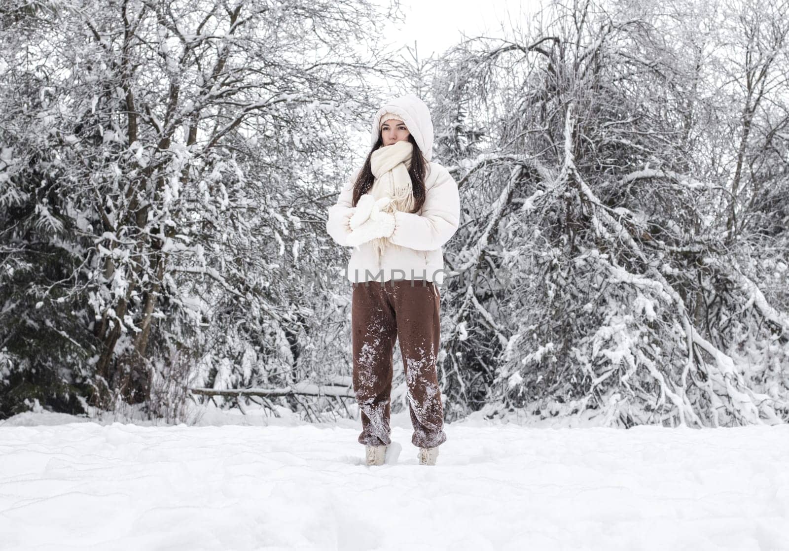 Portrait of a beautiful young caucasian brunette girl in a white down jacket with a hood, brown pants and knitted mittens standing alone and thoughtfully on a snowy meadow in a winter forest reserve in belgium, close-up side view.Concept winter vacation.