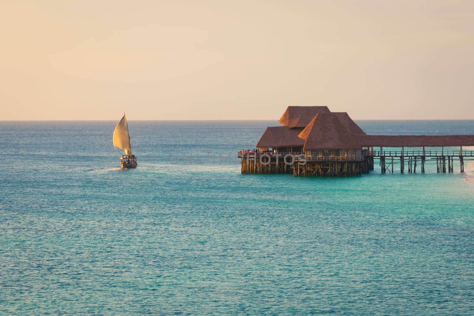 Dhow boat sails in the ocean near beautiful thatch stilt house restaurant at sunset by Robertobinetti70