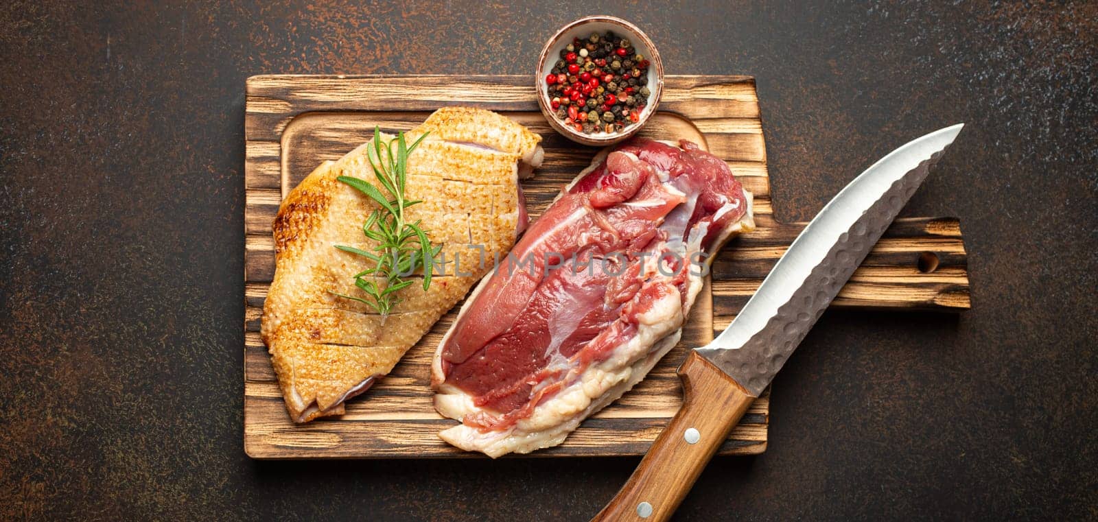 Two raw uncooked duck breast fillets with skin, seasoned with salt, pepper, rosemary top view on wooden cutting board with knife, dark brown concrete rustic background.