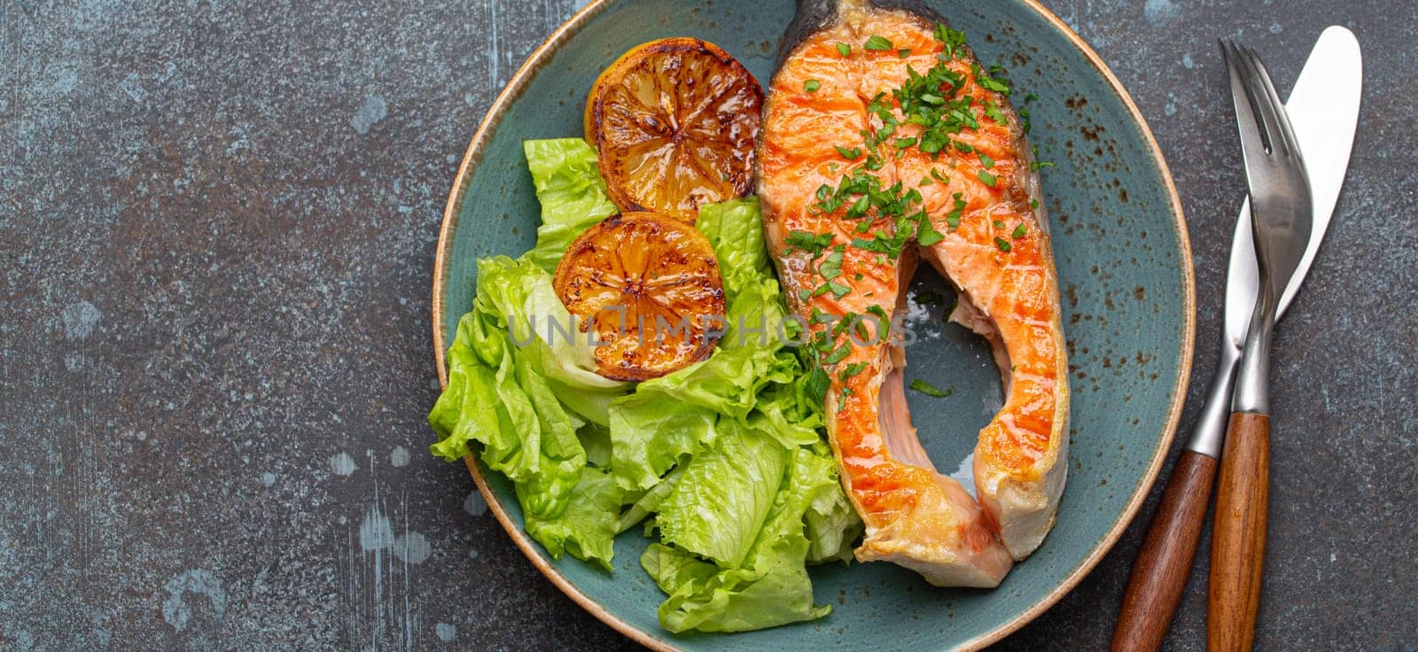 Grilled fish salmon steak and green salad with lemon on ceramic plate on rustic blue stone background top view, balanced diet or healthy nutrition meal with salmon and veggies, space for text.