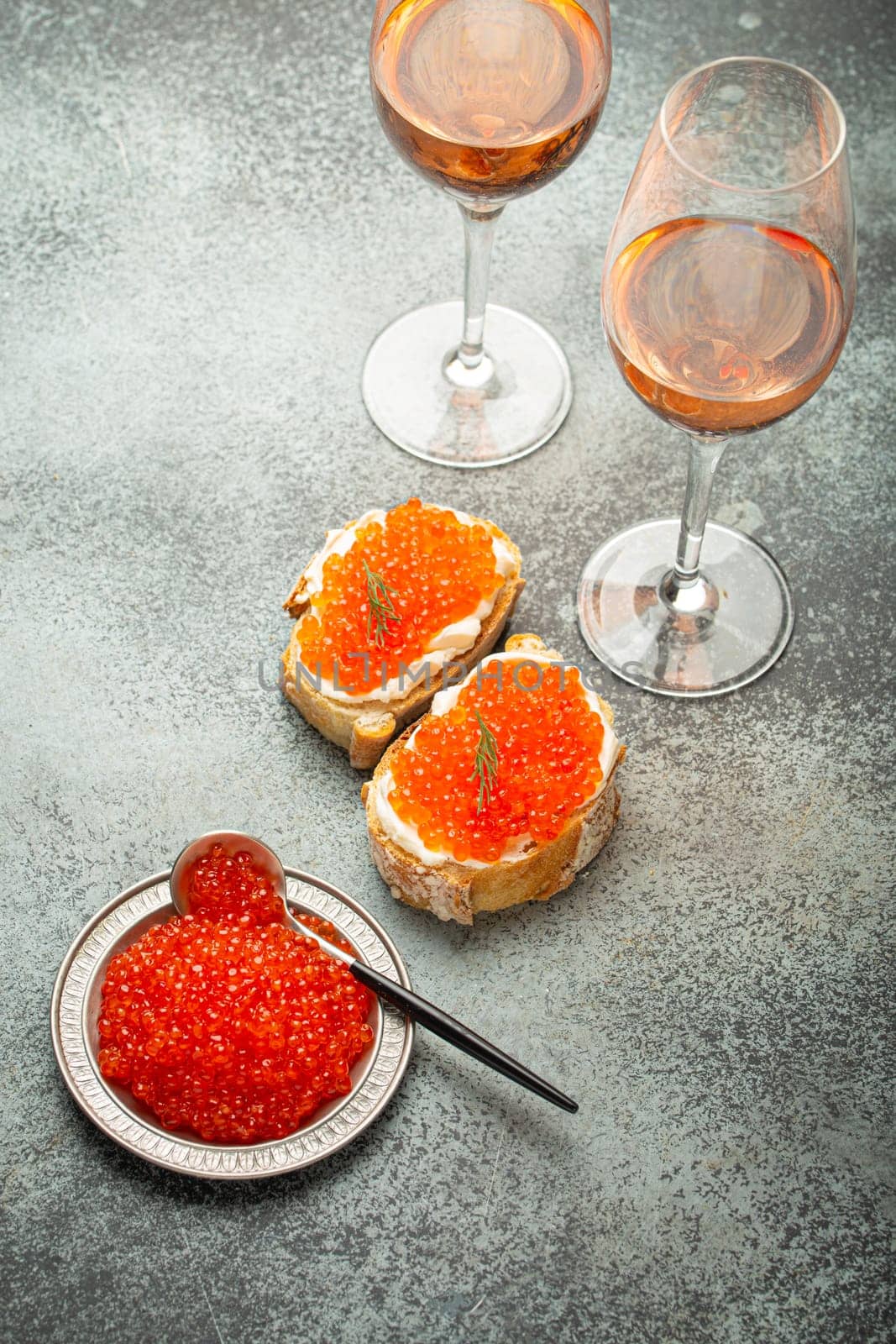 Small metal plate with red salmon caviar, two caviar toasts canapé, two glasses of champagne top view on grey concrete background, festive luxury delicacy and appetizer.