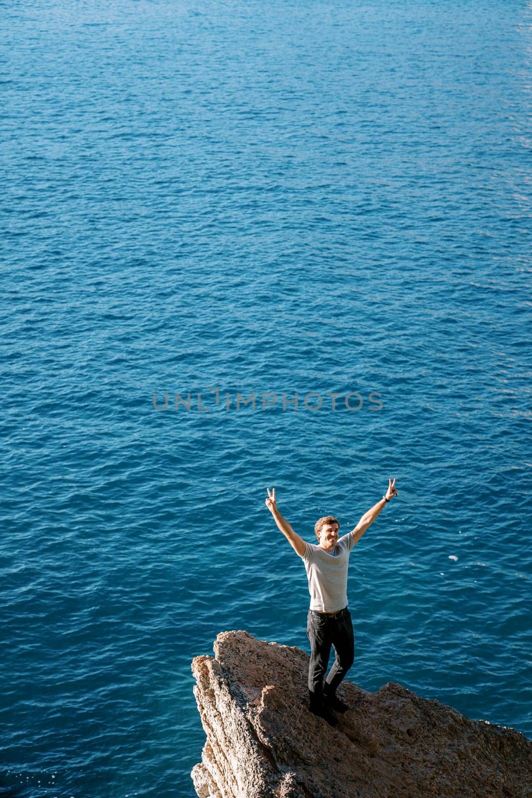 Smiling young man stands on a stone ledge above the sea with his hands up by Nadtochiy