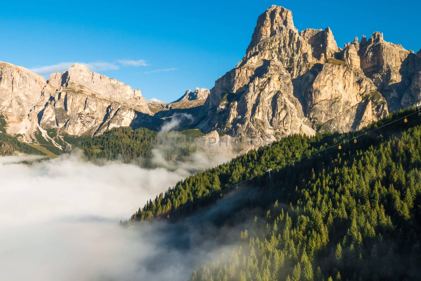 Rocky peak of giant Alps rises against clear blue sky at sunrise. Dense fog envelops high mountains in a captivating aerial view on a sunny summer morning