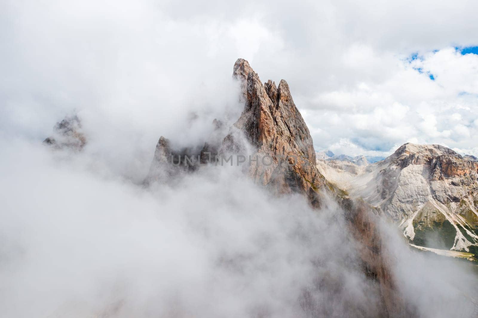 Dense fog descends onto Seceda mountain peaks on sunny summer day. Giant ridgeline with bare peak and steep slope in Italian Alps aerial view