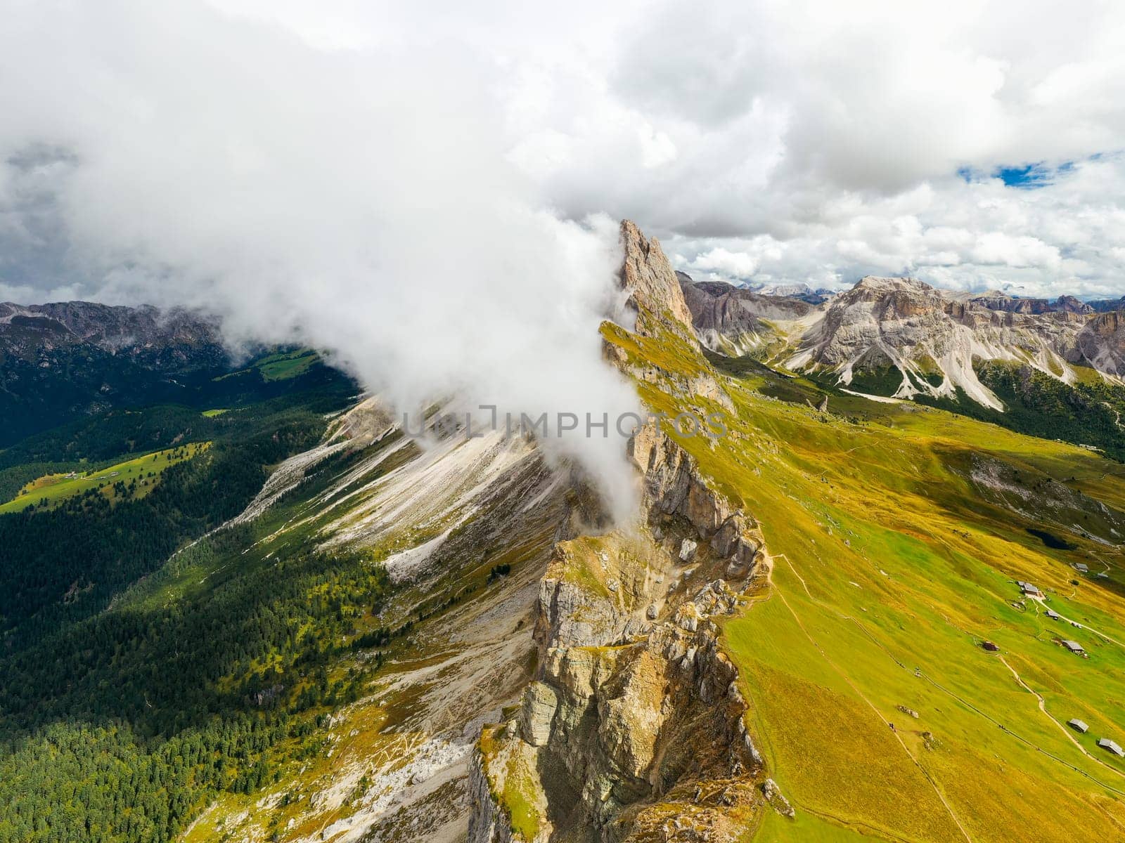 White heavy clouds descend covering Seceda bare peaks of mountain range. Scenic Italian Alps and dense fog in summer morning aerial view