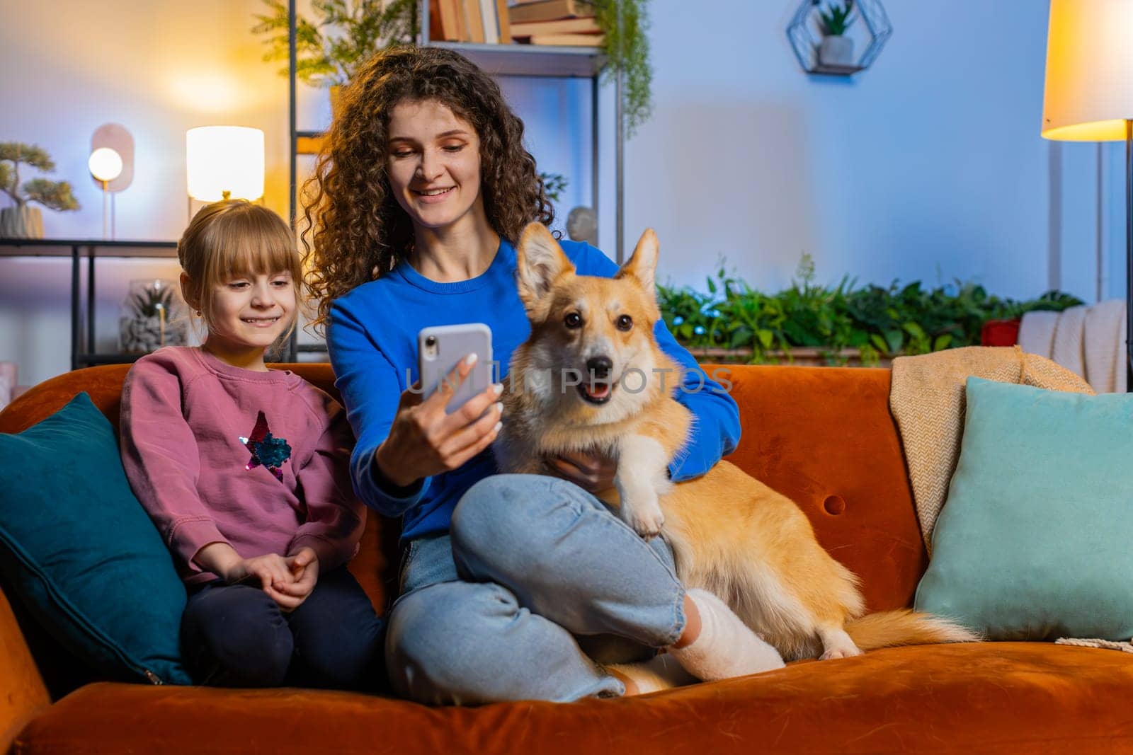 Cheerful mother and daughter child girl taking selfie with corgi dog using smartphone on sofa. Happy family sharing social media online video call sits on sofa in living room at home during weekends