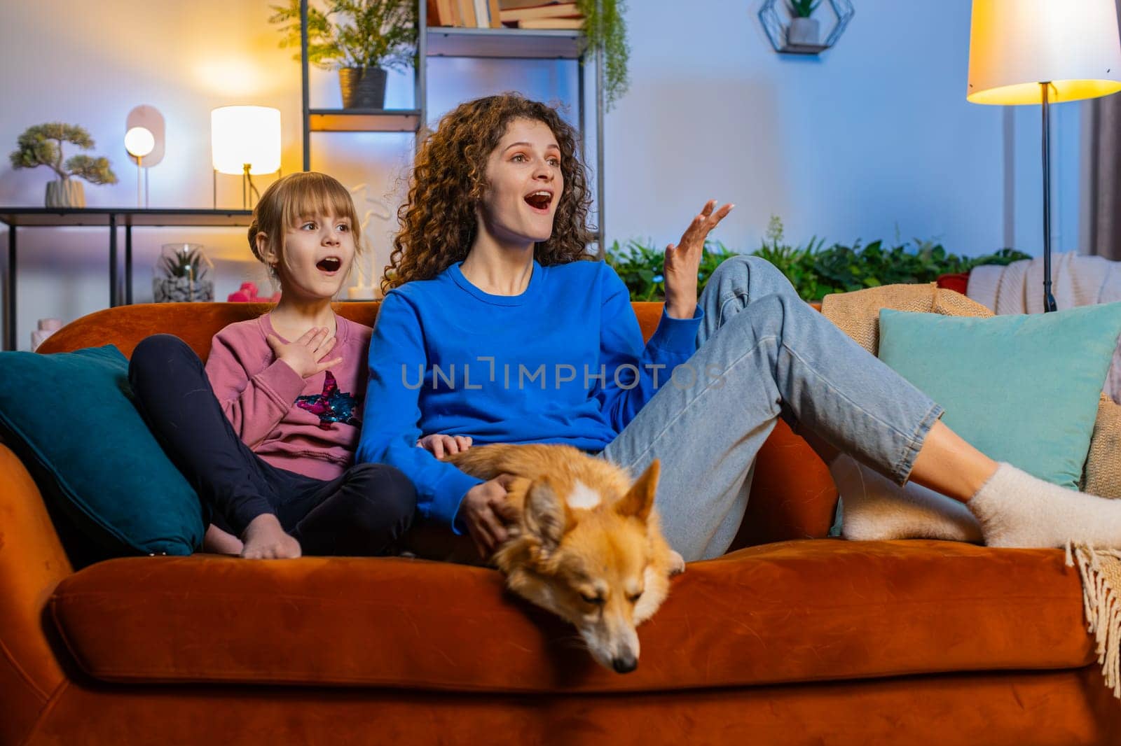 Surprised mother and daughter child girl sitting beside corgi dog watching TV in night room at home. Happy family in casual clothes watching favorite TV show sits on sofa in apartment during weekend.