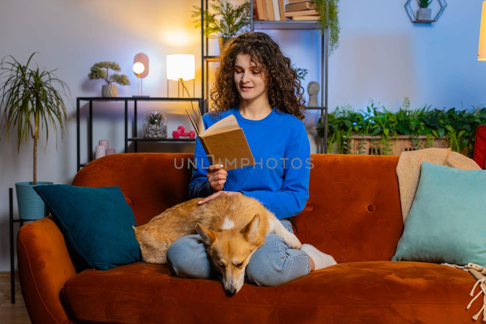 Beautiful pretty woman with curly hair in casual clothes reading book while sitting beside cute corgi dog on sofa in living room at home. Happy female girl spending leisure time with pet dog on couch.