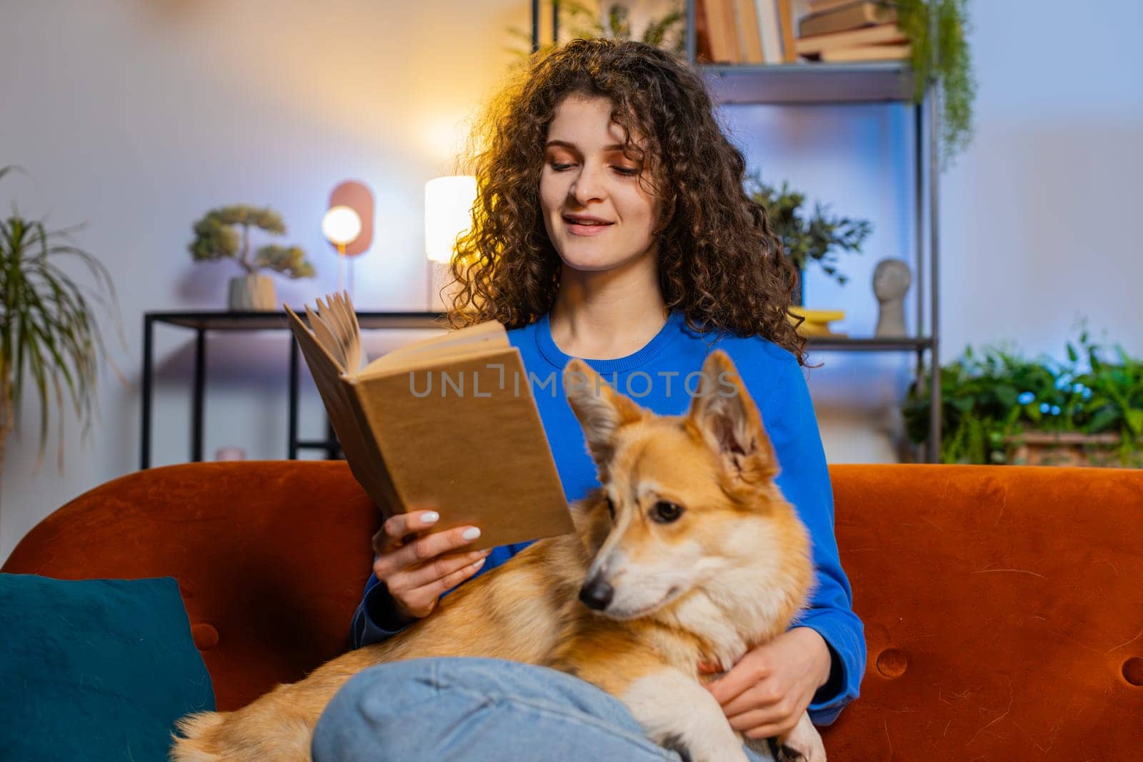 Beautiful young woman with curly hair in casual clothes reading book while sitting beside cute corgi dog on sofa in living room at home. Happy female girl spending leisure time with pet dog on sofa