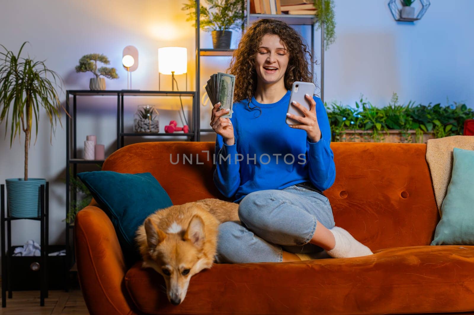 Successful rich woman holding smartphone and waving dollar bill money fan sitting on sofa beside corgi dog at home. Happy girl winning online lottery game planning vacation calculating budget concept