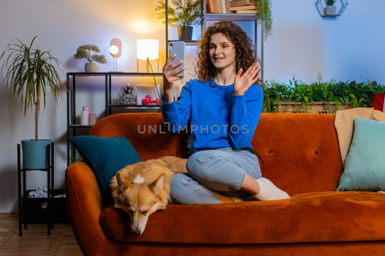 Caucasian young woman taking selfie on smartphone waving hello communicating video call online with subscribers, recording stories vlog for social media. Female girl sitting at home with pet corgi dog