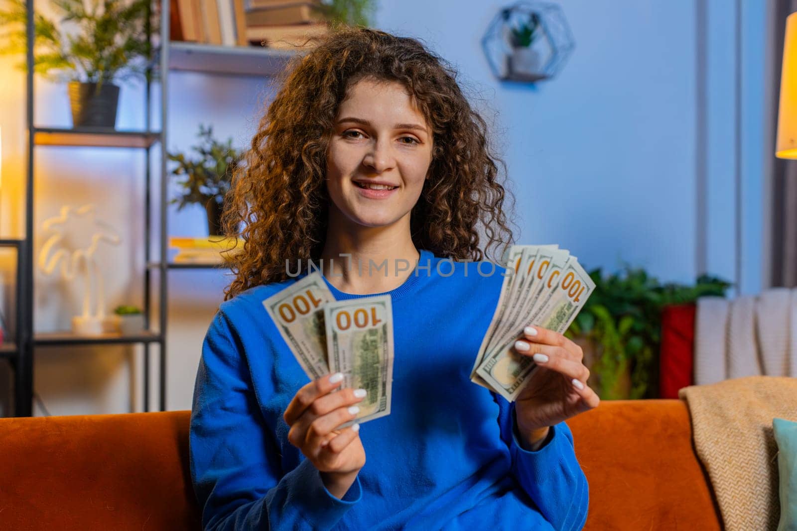 Successful happy rich woman with curly hair counting dollar banknotes while sitting on couch in living room at home. Smiling girl calculating budget, savings, earnings, planning vacation in apartment.