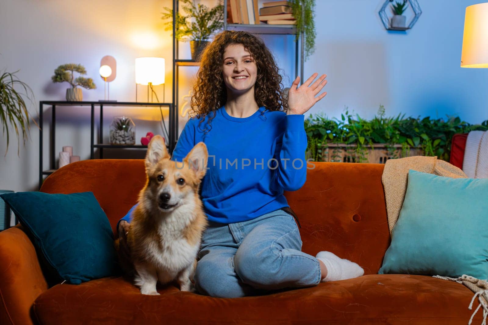 Portrait of smiling young woman with curly hair waving hands saying hello and looking at camera. Cheerful beautiful lady girl in casual clothes greeting while sitting on sofa in living room at home.
