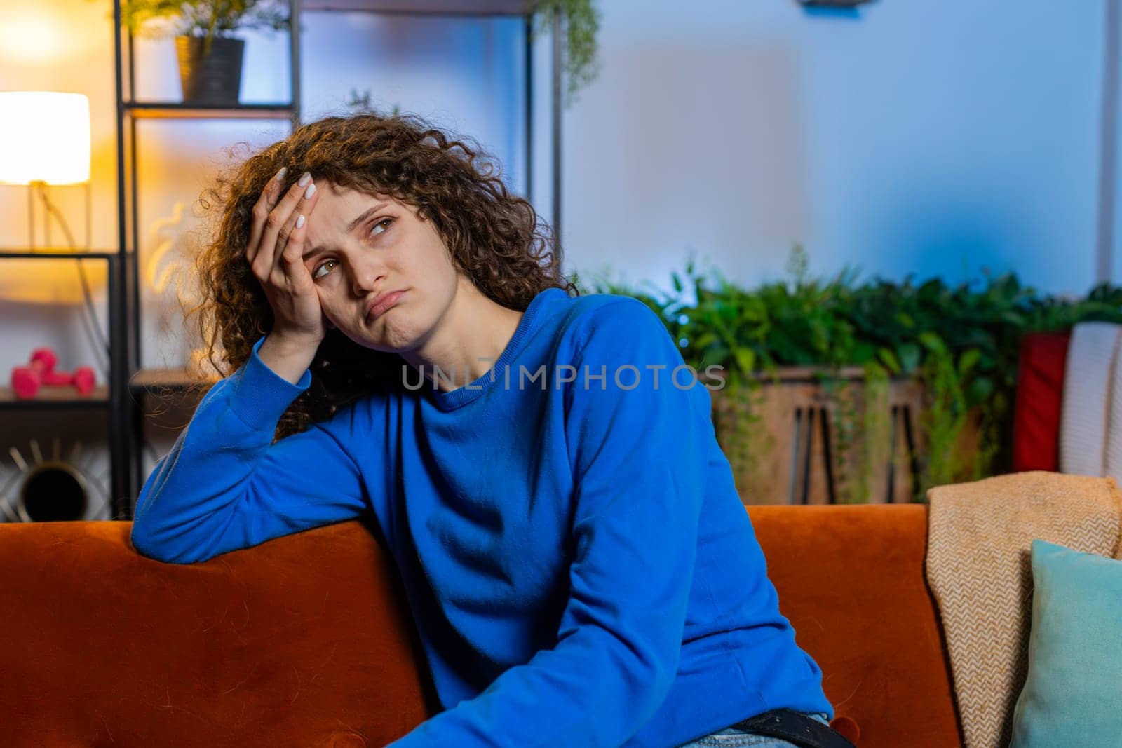 Upset lonely young woman with curly hair in casual clothes thinking about problems sitting on sofa in living room at home. Bored female girl feeling bad annoyed burnout while looking away in apartment