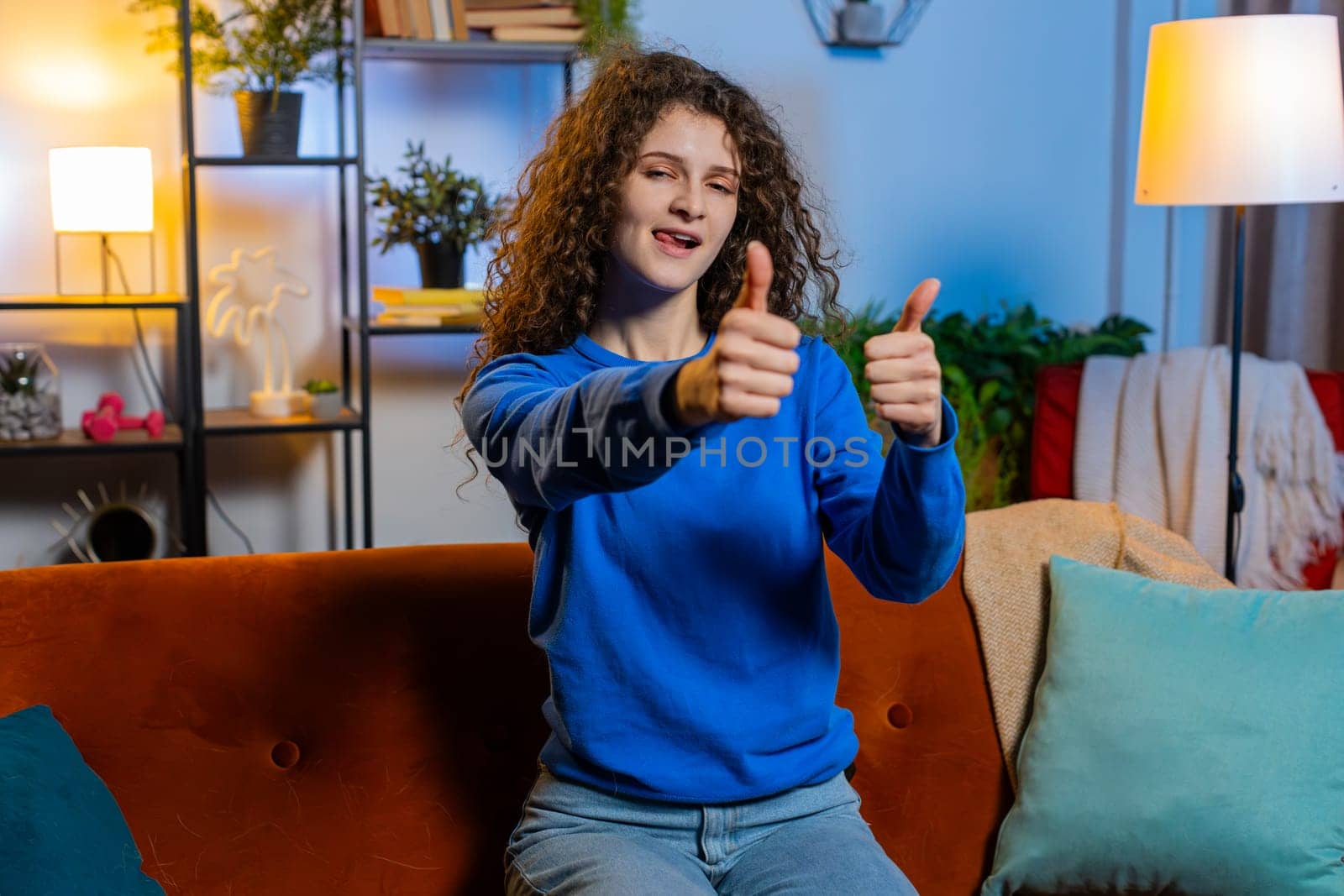 Portrait of smiling young happy woman with curly hair showing thumbs up with hands sitting on sofa in living room. Successful female girl gesturing like positive approve and looking at camera at home