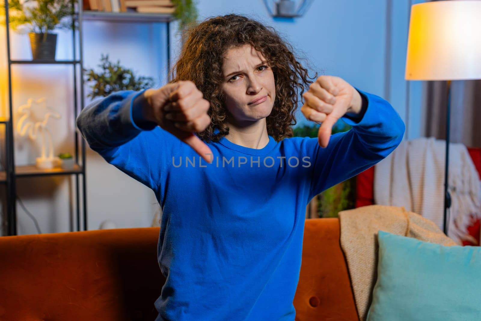 Portrait of upset young woman with curly hair showing thumbs down dislike gesture sitting on couch in living room. Displeased Caucasian female adult girl expressing discontent and disapproval at home