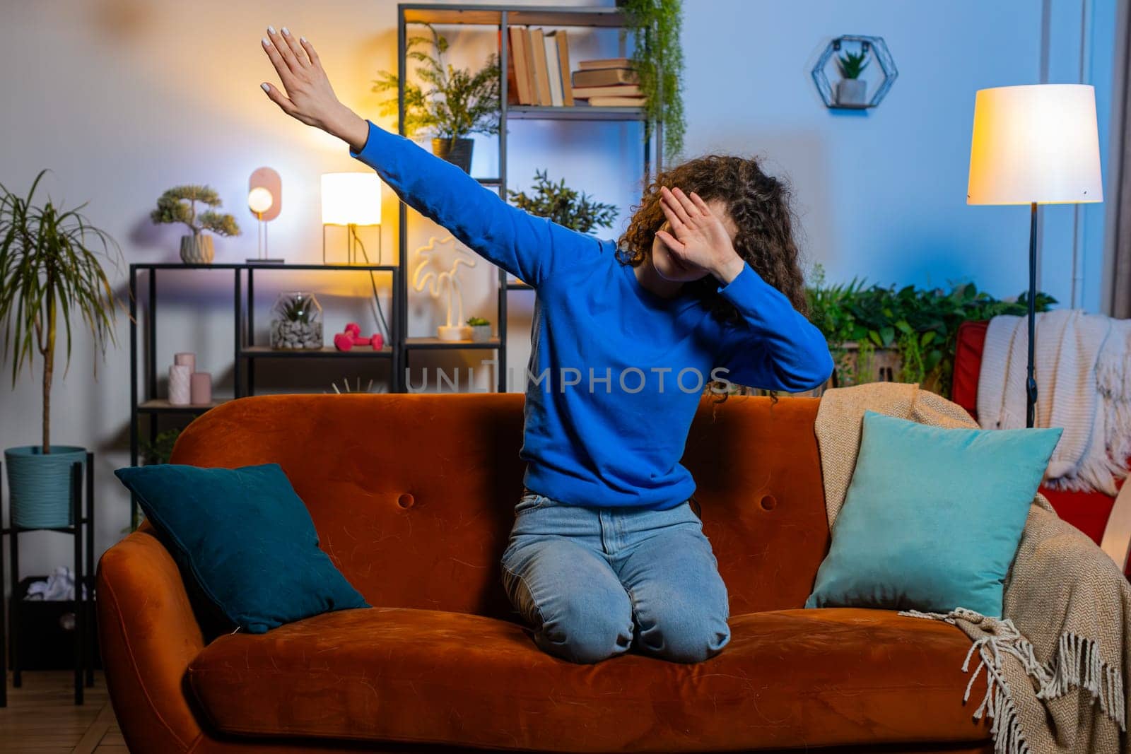 Excited young woman with curly hair having fun dancing and moving to rhythm sitting on sofa in living room. Cheerful female girl making dub dance gesture in apartment. Trendy girl dabbing at home.
