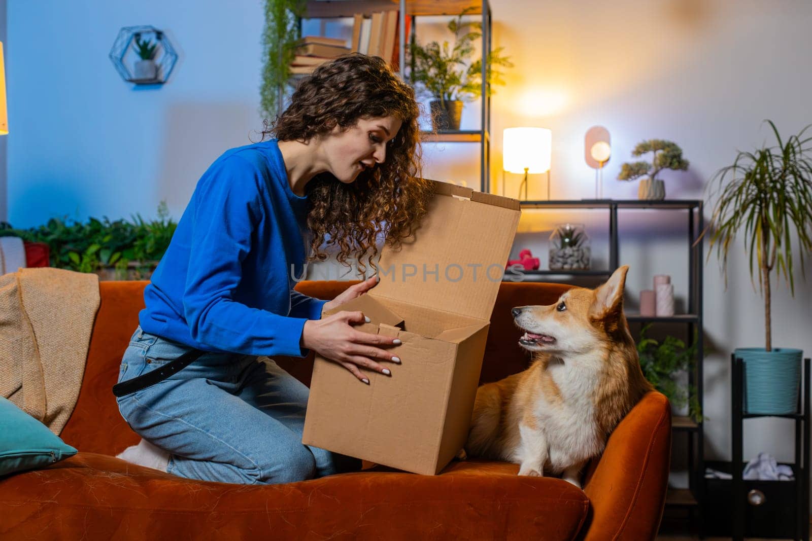 Shocked satisfied woman unpacking delivery parcel. Girl shopper online shop customer opening cardboard box receiving purchase gift by fast postal shipping beside cute corgi dog in living room at home