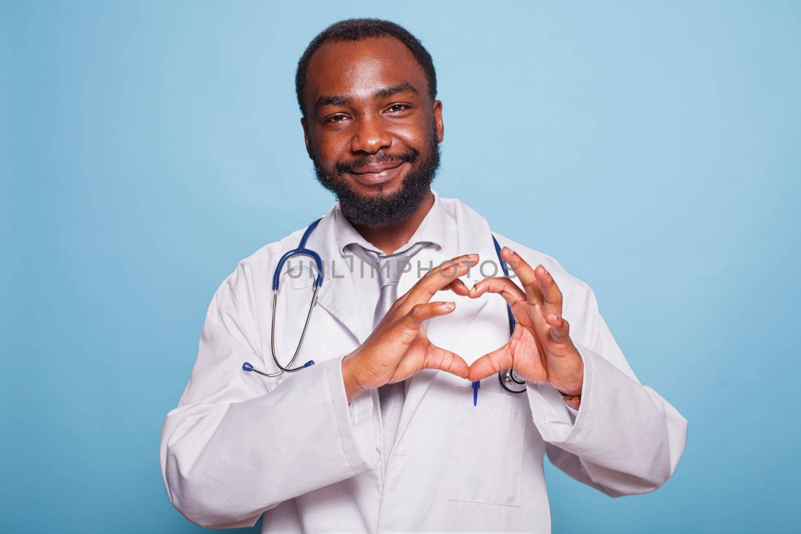 African American medical practitioner with a stethoscope standing over an isolated blue background smiling and showing a heart symbol with hands. Romantic concept.