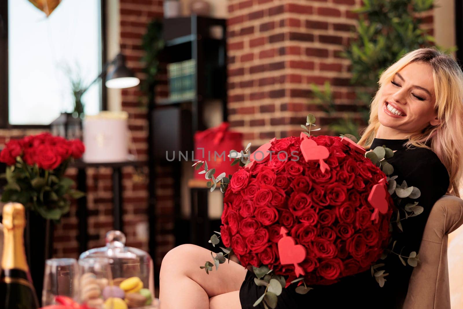 Girlfriend standing in living room filled with romantic gifts, holding red roses large bouquet. Beautiful blonde woman in elegant black dress enjoying celebrating valentine s day. Love concept