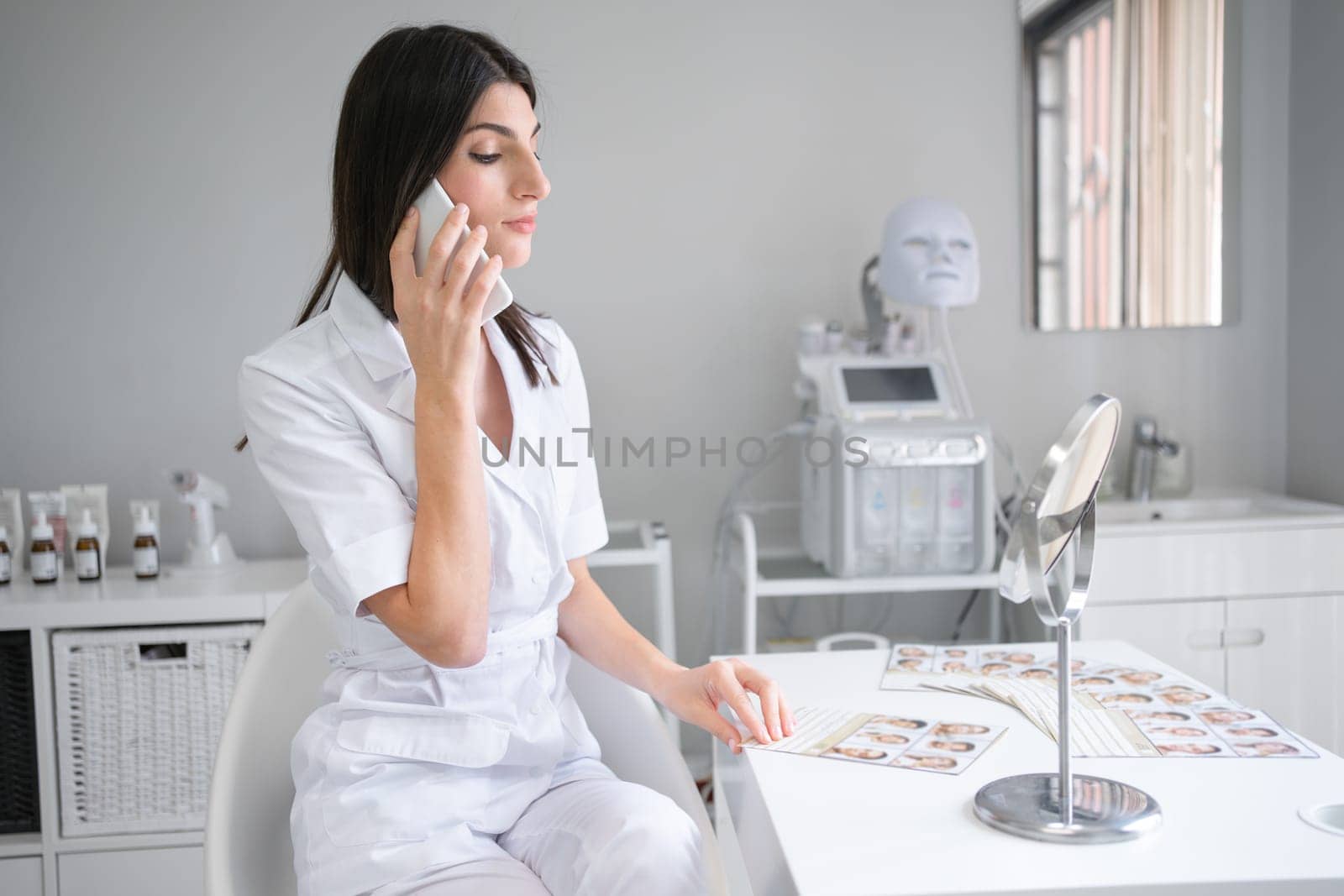 Young female cosmetologist in white uniform talking on smartphone at desk. Woman beautician discussing facial skin treatments for clients on phone call at beauty spa salon.