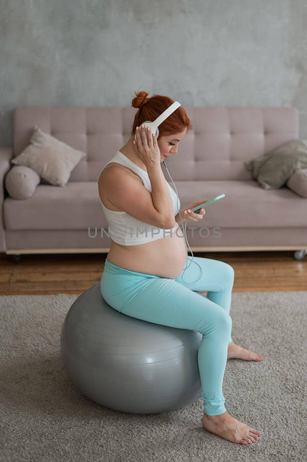 Caucasian red-haired pregnant woman sitting on fitball and listening to music with headphones at home