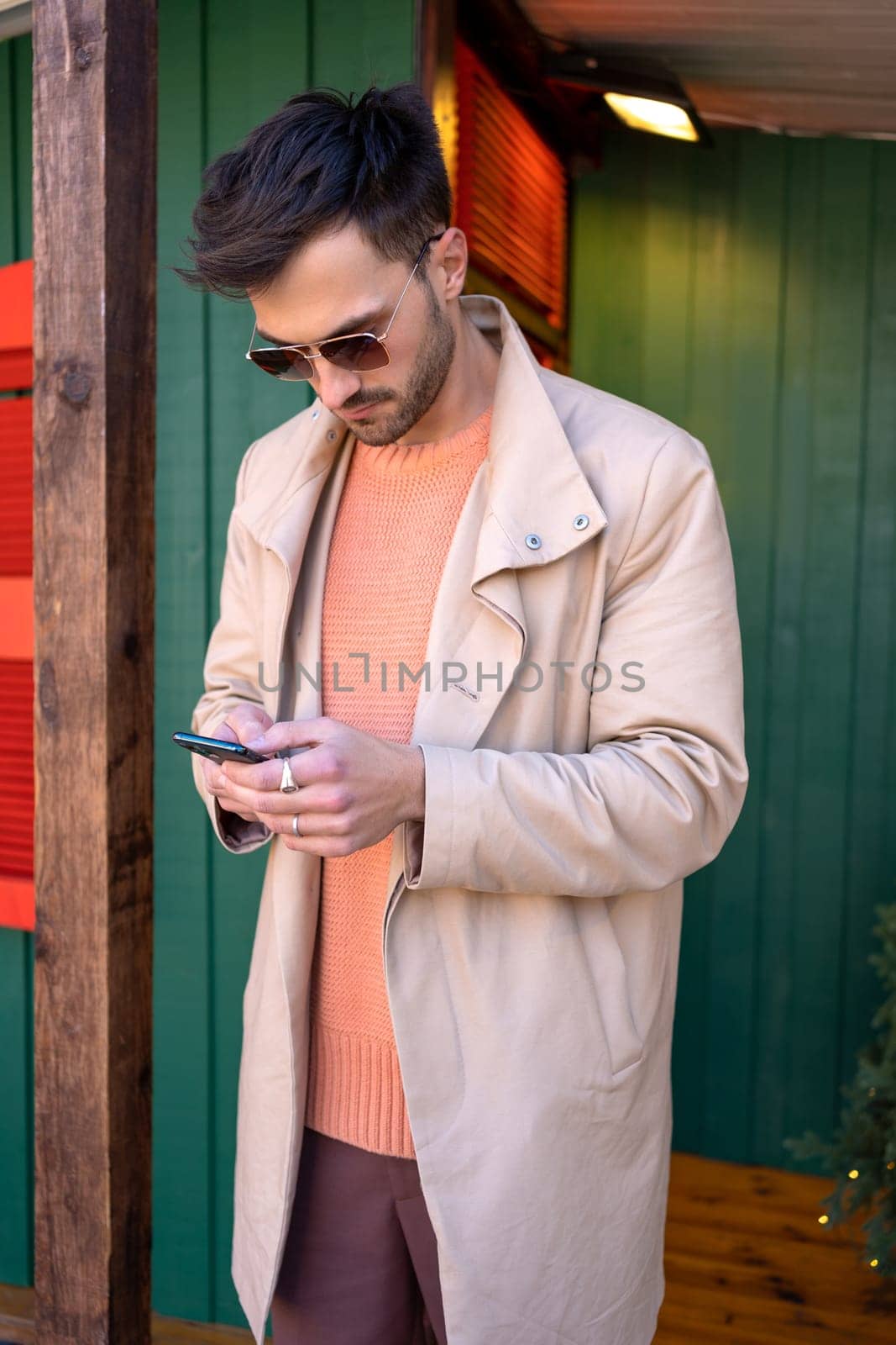 Handsome man in coat using smartphone near wooden house by andreonegin