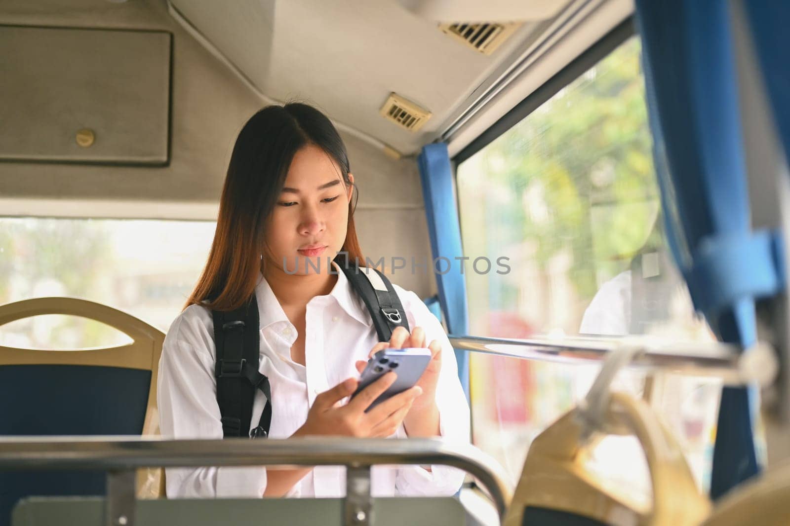 Young woman with backpack using mobile phone while traveling in bus on sunny day by prathanchorruangsak