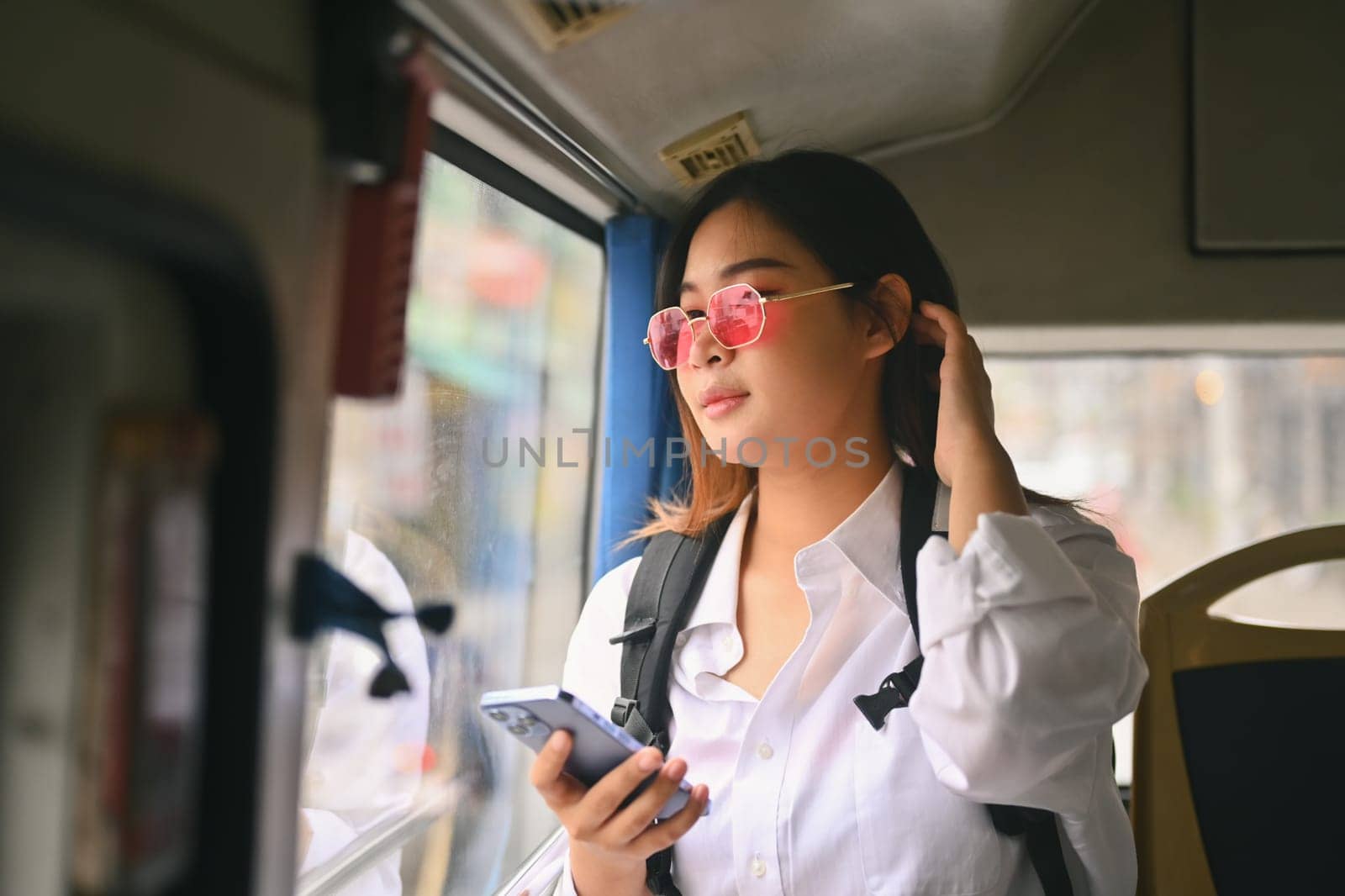 Stylish female tourist in pink glasses using mobile phone while traveling by bus. Public transportation concept.