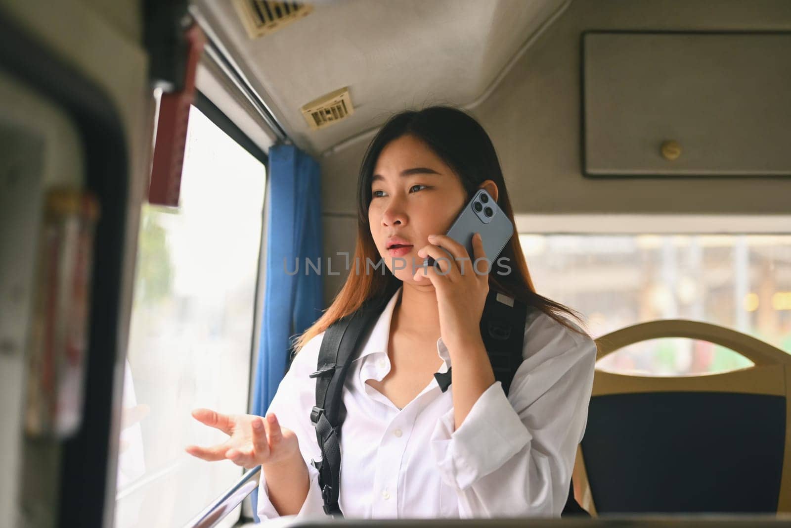 Young Asian woman with backpack talking on mobile phone inside public transport bus. by prathanchorruangsak