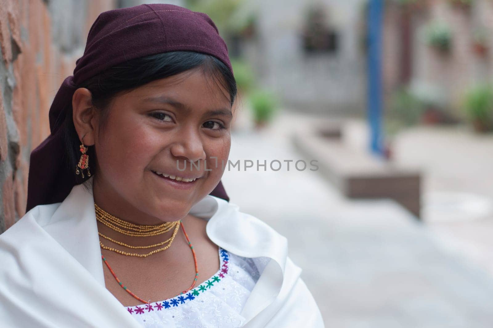 A young indigenous girl wearing traditional dress including a headscarf and jewelry smiles warmly while leaning against a brick wall in a cobblestone alley. by Raulmartin